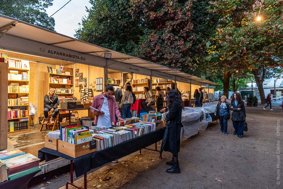 Book stalls lit up at dusk at the Feira do Livro do Porto, with visitors browsing the shelves under the trees