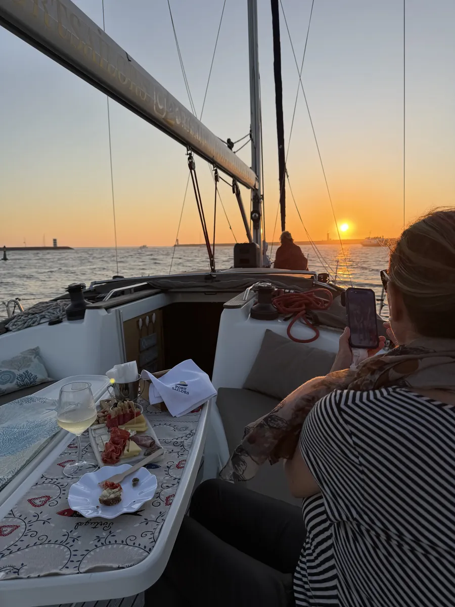 Guests on the deck of a Port Sailors sailboat at sunset, with a charcuterie board and a glass of Port